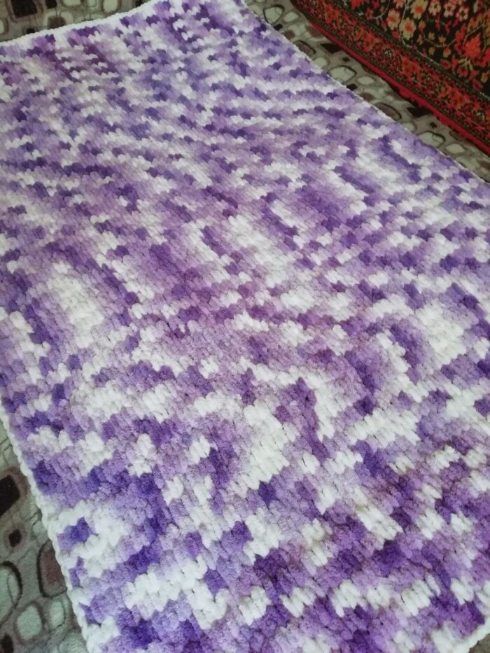 Этот плед выполнен из плюшевой пряжи Alize Puffy производства Турции , что придаёт ему особую мягкость и приятную текстуру. Изделие отличается объёмной вязкой с рельефным узором, создающим ощущение уюта и тепла. Особенности пледа: Цветовая гамма: сочетание нежных оттенков фиолетового и белого. Цвета плавно перетекают друг в друга, создавая эффект акварельного рисунка. Такой дизайн выглядит стильно и свежо, подойдёт для любого интерьера. Структура: вязка выполнена крупными петлями, что придаёт пледу дополнительную пушистость. Рельефный узор напоминает «петельчатую» технику, характерную для пряжи Puffy. Форма: прямоугольная, классическая форма пледа. Края аккуратно обработаны, без дополнительной каймы — акцент сделан на основной узор. Тактильные ощущения: благодаря плюшевой пряже плед очень мягкий, приятный на ощупь. Отлично подойдёт для использования в холодное время года — как для укрывания, так и в качестве декоративного элемента. Назначение: универсален — подойдёт для детской комнаты, спальни или гостиной. Можно использовать как покрывало, плед для отдыха или декоративный акцент на диване/кресле. Преимущества: гипоаллергенный материал (если пряжа акриловая); простота ухода (обычно такие пледы можно стирать в машинке на деликатном режиме); долговечность за счёт плотной вязки; эстетичный внешний вид — плед выглядит аккуратно и стильно. В целом, это уютный и практичный аксессуар, который добавит тепла и уюта в любой интерьер.