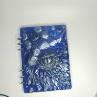 Гримуар «Глаз Дракона»: блокнот, хранящий древние тайны Перед вами не просто блокнот, а магический гримуар — реликвия, рождённая на стыке искусства и волшебства. Его обложка, словно выкованная из драконьей чешуи, переливается глубоким синим цветом, напоминая ночное небо, опалённое дыханием дракона. В центре обложки — **живой драконий глаз**, будто следящий за каждым вашим движением. Этот мистический взгляд пробуждает воображение: кажется, что дракон готов пробудиться в любой момент, чтобы поделиться своими тайнами или защитить записи от посторонних глаз. Рельефные узоры вокруг глаза напоминают чешую и шипы, добавляя артефакту аутентичности. Особенности магического гримуара: * Прочная обложка из эпоксидной смолы с трёхмерным дизайном — не просто украшение, а оберег от злых чар. * Съёмные листы — возможность менять пергамент по своему усмотрению: для записей заклинаний, эскизов мифических существ или тайных посланий. * Глубокий синий цвет — символ мудрости и силы, который настраивает на серьёзный творческий лад. * Металлические кольца — надёжная защита содержимого, как доспехи для рыцаря. Этот блокнот идеально подойдёт: * магам и алхимикам для записи древних формул; * писателям фэнтези для создания эпических историй; * художникам для зарисовок волшебных существ; * искателям приключений, желающим вести дневник своих подвигов. Легенда гласит: тот, кто откроет этот гримуар с чистым сердцем, обретёт вдохновение, способное перенести его в миры, где драконы правят небесами, а магия — часть повседневной жизни. Позвольте «Глазу Дракона» стать вашим проводником в мир фантазии. Откройте обложку — и шагните в сказку!