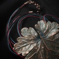 Изысканное колье, центральным элементом которого служит лист садовой герани, выполненный с помощью техники гальванопластика. Лист, окрашенный в нежные оттенки оливкового и болотно-бирюзового, словно парит в воздухе — вощёный шнур искусно выходит из его боковинок, создавая иллюзию невесомости и лёгкого парения.