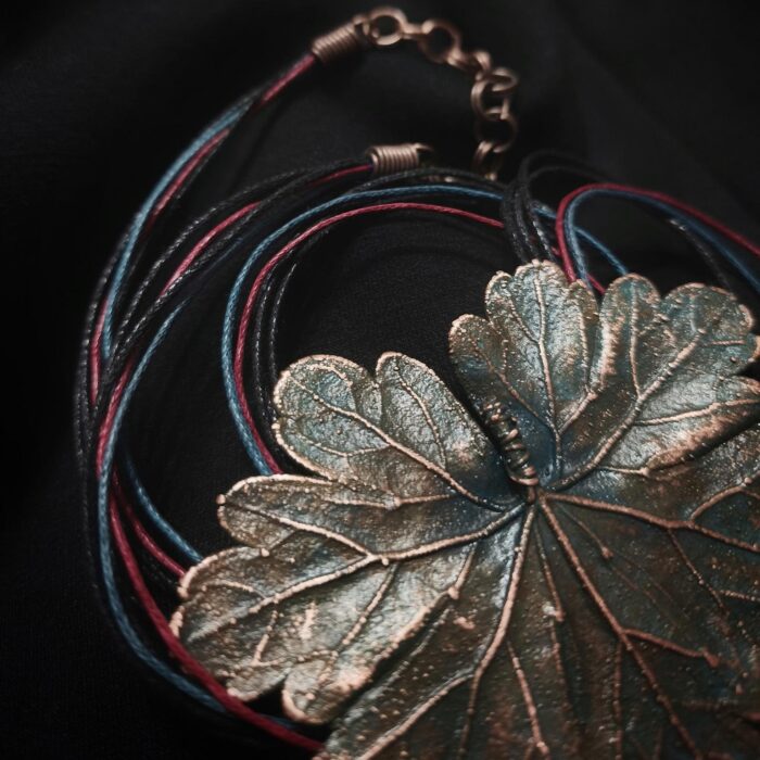 Изысканное колье, центральным элементом которого служит лист садовой герани, выполненный с помощью техники гальванопластика. Лист, окрашенный в нежные оттенки оливкового и болотно-бирюзового, словно парит в воздухе — вощёный шнур искусно выходит из его боковинок, создавая иллюзию невесомости и лёгкого парения.