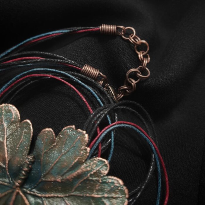 Изысканное колье, центральным элементом которого служит лист садовой герани, выполненный с помощью техники гальванопластика. Лист, окрашенный в нежные оттенки оливкового и болотно-бирюзового, словно парит в воздухе — вощёный шнур искусно выходит из его боковинок, создавая иллюзию невесомости и лёгкого парения.