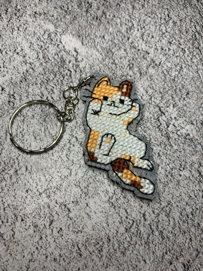 Представляем очаровательный брелок, выполненный в технике вышивки крестиком на пластиковой канве. Центральным элементом дизайна стал расслабленный котик в характерной «ленивой» позе — лежащий на боку с поднятой лапкой. Этот аксессуар сочетает в себе игривость и уют, станет стильным дополнением к ключам, рюкзаку или сумке.