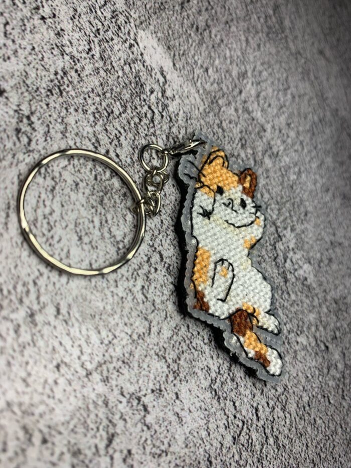 Представляем очаровательный брелок, выполненный в технике вышивки крестиком на пластиковой канве. Центральным элементом дизайна стал расслабленный котик в характерной «ленивой» позе — лежащий на боку с поднятой лапкой. Этот аксессуар сочетает в себе игривость и уют, станет стильным дополнением к ключам, рюкзаку или сумке.