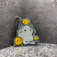 Представляем очаровательный брелок, выполненный в технике вышивки крестиком на пластиковой канве. Центральным элементом дизайна стал милый котик с расслабленным выражением мордочки, украшенный тремя ярко-жёлтыми цыплятами — один на голове и по одному с каждой стороны. Этот аксессуар сочетает в себе игривость и уют, станет стильным дополнением к ключам, рюкзаку или сумке.