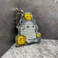 Представляем очаровательный брелок, выполненный в технике вышивки крестиком на пластиковой канве. Центральным элементом дизайна стал милый котик с расслабленным выражением мордочки, украшенный тремя ярко-жёлтыми цыплятами — один на голове и по одному с каждой стороны. Этот аксессуар сочетает в себе игривость и уют, станет стильным дополнением к ключам, рюкзаку или сумке.