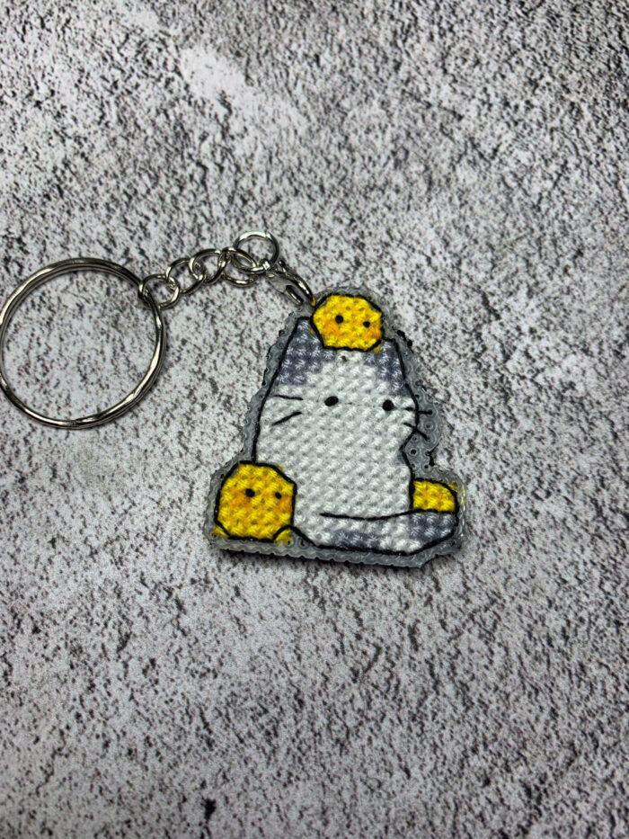 Представляем очаровательный брелок, выполненный в технике вышивки крестиком на пластиковой канве. Центральным элементом дизайна стал милый котик с расслабленным выражением мордочки, украшенный тремя ярко-жёлтыми цыплятами — один на голове и по одному с каждой стороны. Этот аксессуар сочетает в себе игривость и уют, станет стильным дополнением к ключам, рюкзаку или сумке.