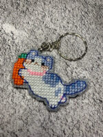 Представляем очаровательный брелок, выполненный в технике вышивки крестиком на пластиковой канве. Центральным элементом дизайна стал милый котик, который держит в лапках ярко-оранжевую морковку с зелёной ботвой. Этот аксессуар сочетает в себе игривость и утончённость, станет стильным дополнением к ключам, рюкзаку или сумке