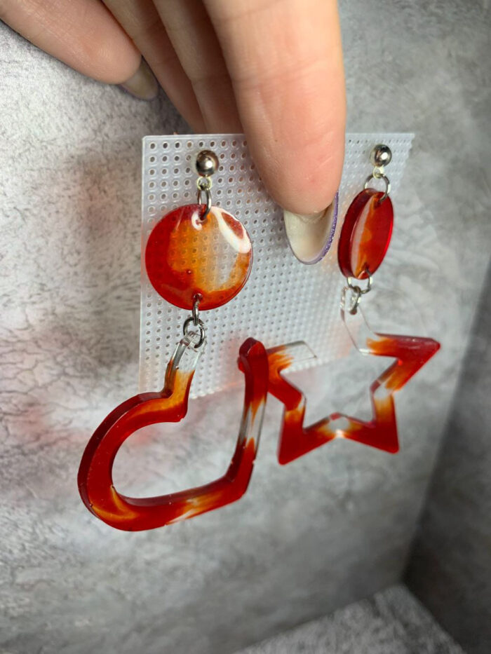 Представляем эффектные серьги из эпоксидной смолы, которые станут ярким акцентом в любом образе! Эта пара сочетает в себе два выразительных элемента: округлый диск и крупную геометрическую фигуру — сердце и звезду. Насыщенная красно-оранжевая гамма с полупрозрачными переливами создаёт ощущение огненного закатного неба, а плавные переходы оттенков придают украшению глубину и объём.