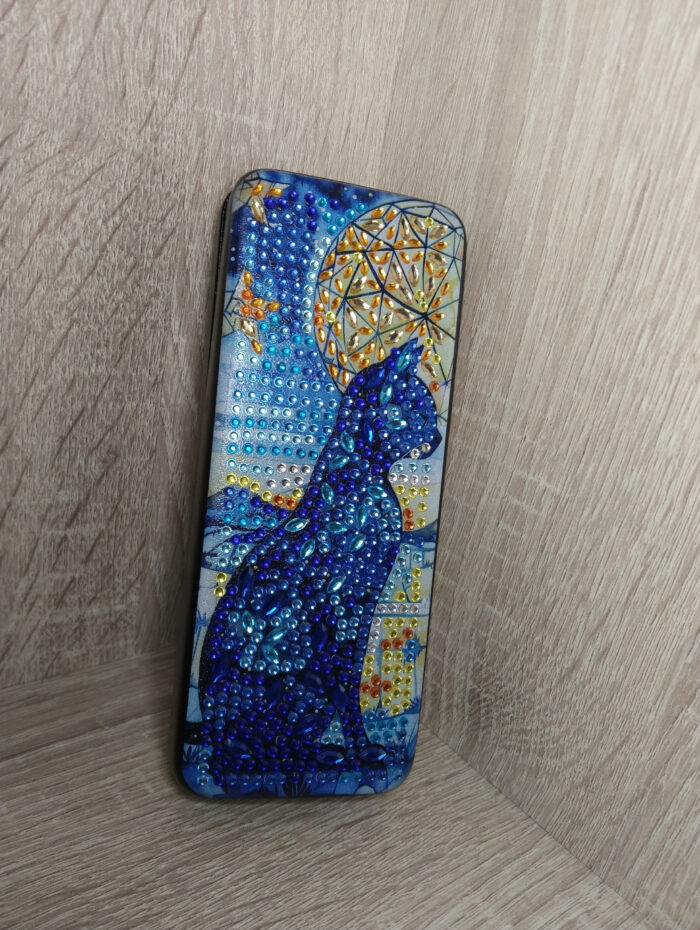 Роскошный футляр для очков, украшенный авторской алмазной вышивкой, превратит хранение аксессуаров в настоящее эстетическое удовольствие. Центральным элементом дизайна стал загадочный силуэт кошки, выполненный с использованием страз насыщенных синих, фиолетовых и бирюзовых оттенков.