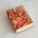 «От сердца к сердцу — через подарок». На фотографии изображена декоративная шкатулка, выполненная в форме закрытой книги с состаренными страницами. Украшена ярким осенним принтом с изображением дубовых и кленовых листьев, ягод и желудей на деревянном фоне. Выполнена из высокопрочного гипса. Изделие оформлено в технике декупаж с элементами винтажного стиля и осенней тематики. Может использоваться для хранения украшений, мелочей, ключей или как интерьерный декор.